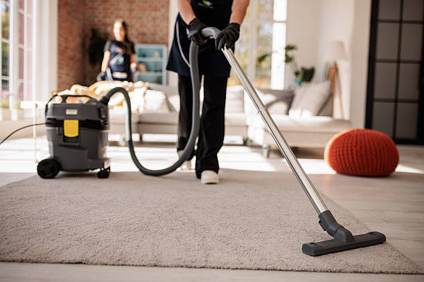 Two female professional cleaners working together to clean a bright, modern apartment. One woman is vacuuming the carpet.